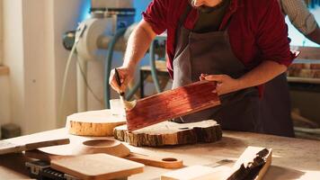 Carpenter in assembly shop starting restoration project on lumber block, applying lacquer on surface. Woodworking specialist coloring wood plank with paintbrush, doing handwork in studio, camera A photo