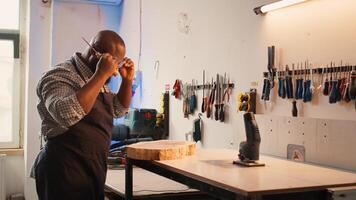 Carpenter inspecting wood in assembling shop, putting safety glasses on and picking tool from rack. Woodworking specialist in studio picking lumber block and carving gear for it, camera B photo