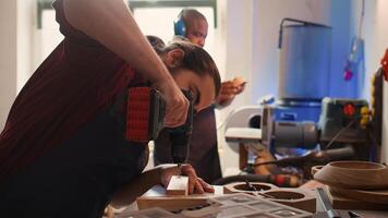 Cabinetmaker drills hole into lumber block, doing furniture assembling in studio, close up. Manufacturer in assembly shop inserting nail into piece of wood using electric power drill, camera B photo