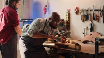 Sculptor shaping raw timber using chisel and hammer in carpentry shop with coworker cleaning workbench in background. BIPOC man making wood sculptures, engraving plank with tools, camera A photo
