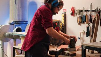 Carpenter at work consulting blueprint, using orbital sander on lumber, wearing safety glasses. Artisan with protecting equipment uses angle grinder on wood after checking schematics, camera A photo