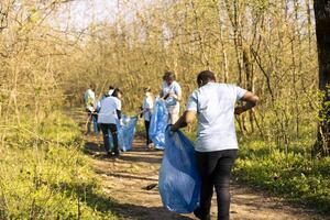 African american woman grabbing junk and plastic waste from the forest, doing voluntary work to preserve and protect the environment. Female activist using claw tool to collect garbage and junk. photo