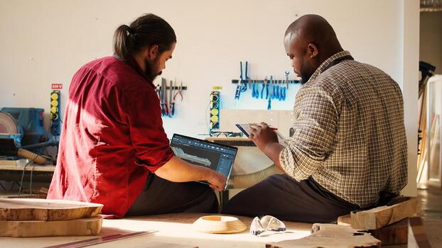 BIPOC artisan and apprentice comparing wood art piece schematic with CAD software 3D render on laptop. Woodworking experts in studio crosscheck drawing on clipboard with notebook blueprint, camera B photo