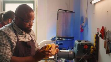 BIPOC man in studio with woodworking tools in background using sandpaper for sanding wooden surface. Woodworking expert using abrasive sponge to fix damages suffered by piece of wood, camera B photo