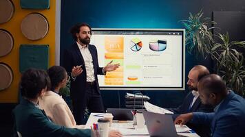 Team leader showing infographics data on interactive board, gathering with workers in a briefing meeting presentation. Colleagues study the reports to adapt to evolving market conditions. Camera B. photo