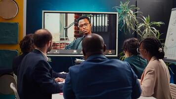 Management team meeting with CEO on videocall connection in a boardroom, briefing him about market trends and project resource allocation. Employees discuss with the director. Camera B. photo