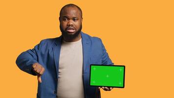 Upset african american man shows thumbs down sign gesturing holding isolated screen tablet, disapproving. BIPOC person does rejection hand gesture regarding mockup device, studio background, camera A photo