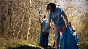 Young volunteer doing litter cleanup with a long claw to grab trash and store it in a garbage bag, dealing with pollution an illegal dumping problem. Activist protecting natural ecosystem. Camera B. photo