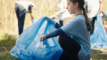 Tired little girl collecting trash and plastic bottles from the forest, helping the volunteers with litter cleanup. Child activist picking up rubbish in a garbage disposal bag. Close up. Camera A. photo