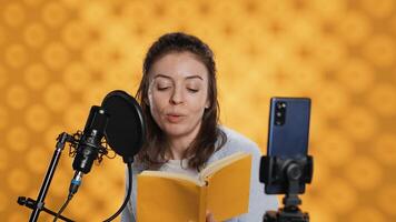 Narrator looking at phone camera, filming herself reading book for kids, creating audiobook, studio background. Content creator doing vlog while producing digital recording of children novel, camera A photo