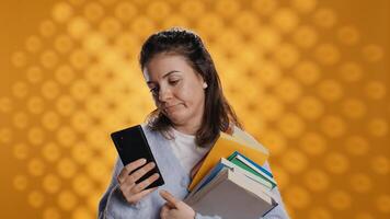 Upset woman holding stack of book, preparing for school exam using phone, studio background. Student with pile of textbooks pouting while researching for university homework using smartphone, camera B photo
