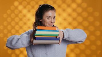Cheerful woman holding stack of books arriving in studio with yellow backdrop, smiling. Joyous person walking with pile of novels, feeling delighted and eager to read, camera B photo