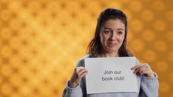Cheerful book club president holds message urging people to join them, talking about importance of lecture, studio background. Smiling woman inviting bookworms to enlist in her association, camera A photo