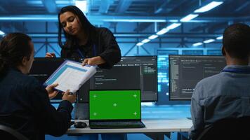Green screen laptop in data center in front of manager and IT support specialists working together, supervising server rows. Team leader and employees overseeing supercomputers using mockup notebook photo