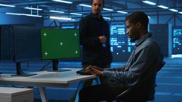 Technician upgrading equipment in data center using isolated screen display. Professional at PC desk in high tech establishment overseeing server rows with chroma key monitor, panning shot photo