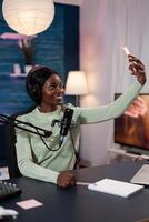 Vlogger woman with headphones smiling while taking selfie using modern smartphone during live stream. Social media creator recording tutorial using broadcasting production equipment. On air talk photo
