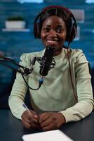 Portrait of smiling infuencer wearing headpones talking into professional microphone answering subscribers question during live stream. Social media vlogger recording content. On air talk show photo