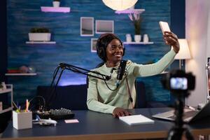 Vlogger woman smiling at camera while taking selfie posting on social media for online content. Influencer person recording podcast vlog using professional filming equipment. On air talk show photo