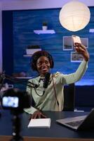 Woman influencer with headphones holding smartphone taking photo while recording vlog content using vlogging production equipment. African american social media vlogger hosting on air talk show