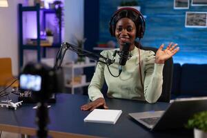 Social media woman creator greeting subscribers while recording content for youtube channel. Influencer sitting in podcast studio filming tutorial using production equipment. On air talk show photo