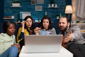 Portrait of multi-ethnic friends sitting on couch watching online movie on laptop computer enjoying hanging out together. Group of people enjoying relaxing time in living room. Friendship concept photo
