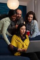 Multi-ethnic friends smiling while watching online comedy series on laptop computer during entertainment movie home night. Group of people enjoying hanging out together. Friendship concept photo