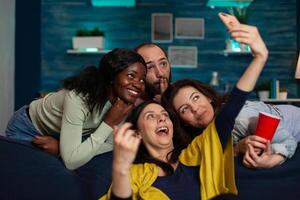 Multi-ethnic friends smiling at camera while smiling woman taking selfie sharing photos on social media celebrating wekeend at home. Group of people making funny expressio. Hanging out concept