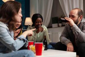 Multi-ethnic group of people enjoying spending time together laughing during wekeend party sitting on sofa in living room. Friends drinking beer, eating snack. Hanging out concept photo