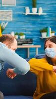 Friends with protection mask socializing during party sitting on sofa touching elbow keeping social distancing greeting each other preventing spread of coronavirus, holding beer bottles in living room photo