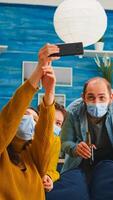 Cheerful happy mixed race friends taking selfie wearing face mask keeping social distancing against spread of virus sitting on couch in living room. Diverse people socializing keeping social distance photo