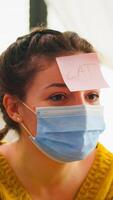 Happy woman gesturing playing name game with multiethnic friends having sticky notes on forehead wearing face mask keeping social distancing. Diverse group of people having fun at new normal party photo