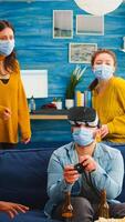 Group of multi ethnic friends having fun playing games using virtual reality headset and joystick wearing face mask keeping social distancing. Diverse people enjoying at new normal party photo