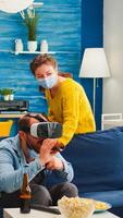 Excited man spending time with friends experiencing playing games with vr headset wearing face mask to prevent coronavirus spread in global pandemic. Diverse people having fun at new normal party photo