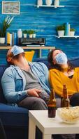 Group of multi ethnic friends watching comedy show on tv laughing wearing face mask to prevent infection with covid 19, during global pandemic having fun sitting on couch keeping social distance photo