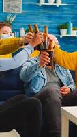 Diverse group of friends watching TV together comedy sitcom eating snacks and drinking beer, clinking bottles and having fun at new normal party, wearing protection face mask against covid 19 virus photo