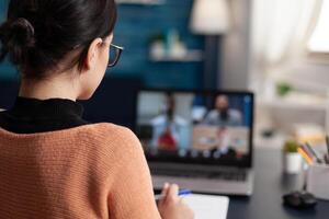 Student in group videocall taking notes. Over the shoulder view of freelancer having briefing call with team in home office and discussing with colleagues in webcam conference. photo