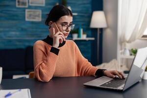 Student at home talking on the phone while using laptop. Programmer with glasses working from home using smartphone and browsing computer. Focused freelancer using technology for remote work. photo