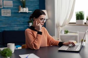 Freelancer in home office working remote using smartphone and laptop. Student with glasses talking on the phone while interacting with computer. Programmer using digital technology for remote job. photo