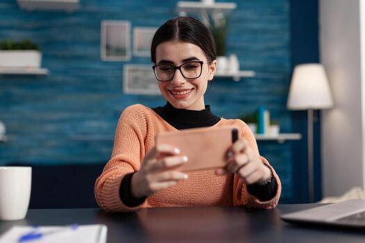 Smiling student with glasses holding smartphone. Game tester relaxing while looking at mobile phone screen. Gamer playing casual online games on smart phone. Freelance app testing. photo