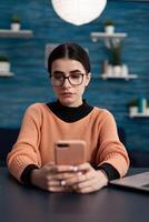 Portrait of student with glasses holding smartphone in hands. Influencer in home office reading email on tochscreen smart phone. Freelancer working remote sitting at desk texting on social media. photo
