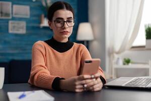 Student with glasses reading email on smartphone. Freelancer holding smart phone looking at content on screen. Influencer sitting at desk browsing social media on tochscreen smart phone. photo