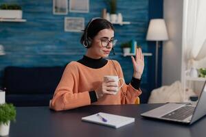 Student with headphones waving hello in laptop videocall. College girl with glasses holding cup attendig online call lecture. Programmer raising hand in remote teleconference course. photo