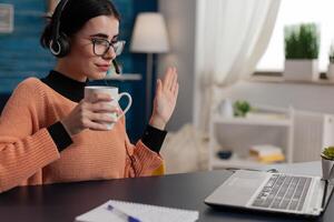 Student with glasses holding cup waving hello at laptop videocall. Programmer with headphones raising hand in remote call lecture. College girl attendig online class raising hand. photo