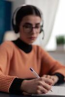 Closeup of student hand with pen taking notes on notepad. College girl with glasses listening to online lecture. Person with headphones writting on notebook in remote course from home office. photo