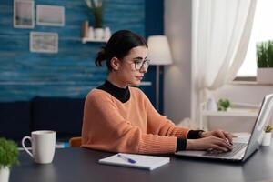 Student with glasses typing on laptop sitting at home desk. College girl with notebook studying remote looking at screen. Freelancer working from home sending email. Programmer browsing the internet. photo