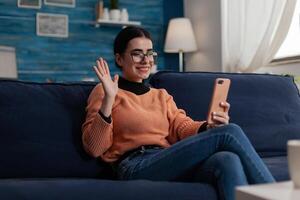 Influencer on couch holding smartphone waves hand at camera. Smiling student with glasses holding mobile phone on sofa in remote videocall. Digital content creator posting on social media. photo