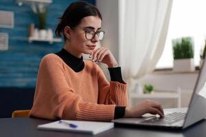 Freelancer with glasses casually typing on laptop keyboard. Student with notebook studying remote smiling at computer screen. Programmer sitting at home desk sending email and browsing the internet. photo