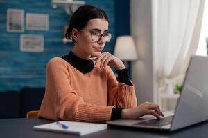 Confident student studying remote looking at laptop screen. Content creator with glasses casually typing sitting at home desk. College girl browsing the internet. Freelancer working from home sending email. photo