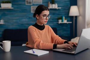 Student with notebook studying remote looking at laptop screen. Influencer with glasses typing on keyboard sitting at home desk. College girl browsing the internet. Freelancer sending email. photo