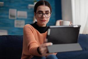 Freelancer with glasses on living room couch holding digital tablet. Student on sofa at home casually looking at gadget display. Content creator in videocall conference while drinking coffee or tea. photo
